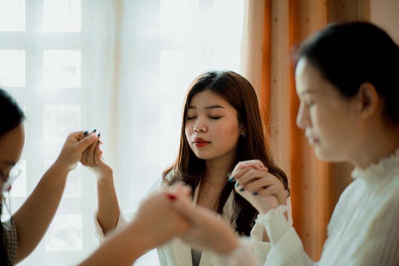 three asian women in a room holding hands with eyes closed praying