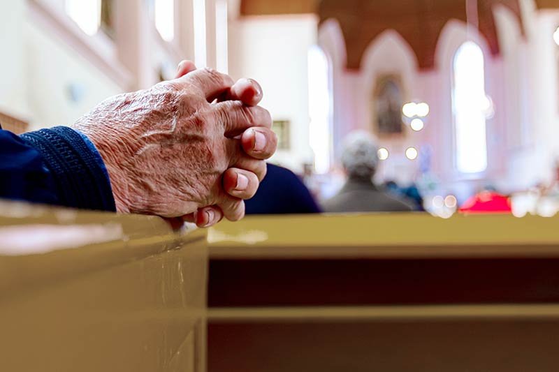 A closeup view of wrinkled elderly hands clasped in prayer resting on a church pew