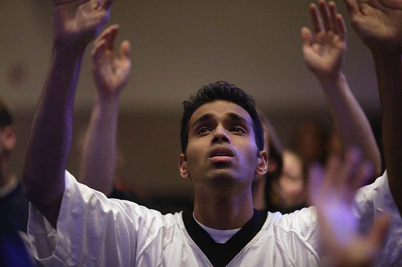 Asian male youth in casual attire with arms raised in praise among a crowd of worshippers